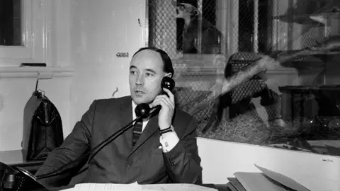 Getty Images A black and white photograph of Desmond Morris, who wears a dark suit. He is holding a rotary phone receiver to his ear, while behind him is a glass display cabinet with a stuffed, small ring-tailed animal within it, posed on a log.