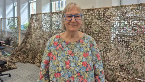 Lindsay Campbell - a woman wearing a floral patterned top, with blue rimmed glasses. She is standing in front of a large green and tan camouflage net. 