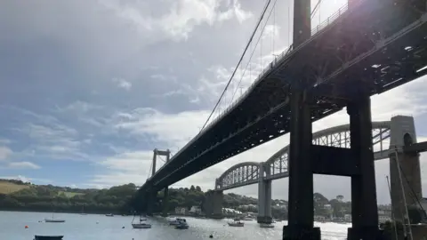 A picture of the Tamar Bridge taken from below. The water can be seen at the bottom of the image and in the distance there are trees and other boats.