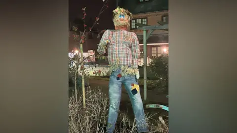 Tex the scarecrow is wearing a tartan shirt and blue jeans with patches on them. He is standing in the middle of a garden arrangement which is in front of buildings.