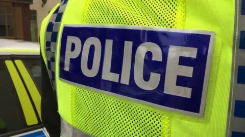 A close-up of a police jacket, which is yellow hi-vis with "police" written on it. A police car is in the background.