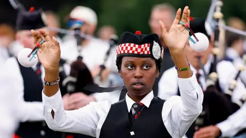 Getty Images A drum major or mace bearer standing in front of a group of bagpipers. She is wearing a traditional piper outfit with hat