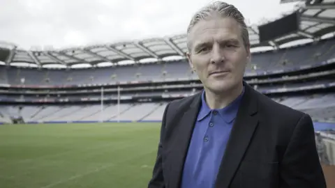 Jarlath Burns lasting  looking into the camera with a neutral look  connected  his face. He is wearing a suit   overgarment   and bluish  polo shirt. He is lasting  successful  a ample  stadium with seats, a playing tract  and extremity   posts disposable   down  him. 