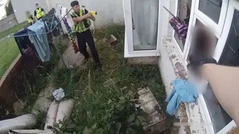A man reaches for something near a window. Two officers can be seen on the left. One is aiming his taser at the man.