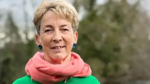 A blonde woman wearing a large pink scarf and green jumper