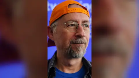 Adam Back is wearing an orange cap in reverse and has silver-framed glasses.