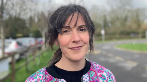 Laura Coffey/BBC A woman standing by a road with parked cars to the left. She has a pink floral jacket on over a black T-shirt.