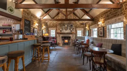 Steve Mortimer The interior of a country pub, with brown tiled flooring and wooden ceiling beams. There is a blue-painted bar with wooden bar stools on one side, and grey booths with small circular tables on the other side, and a wood burner at the far end.