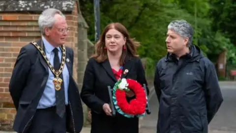 Alex Mayer Alex Mayer with long brown hair, wearing a black coat over a red top and carrying a wreath, is flanked by David Bligh on the left who has white hair and glasses and is wearing a suit with a chain around his neck, and Gennaro Borrelli, who has grey curly hair and is wearing a blue railncoat. They are standing in front of a brick building and trees.