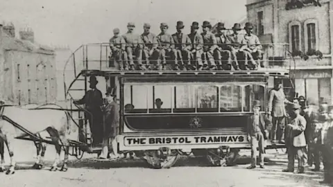 Peter Davey Collection Black and white image of the early trams in use in Bristol. Two horses stand at far left of the picture, at the front of the open-top carriage, with up to twelve people on board. 
