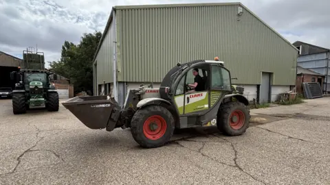 Kian Boyle/BBC A farmyard with large industrial buildings clad in pale green metal sheeting. In the foreground, a telescopic handler with a front bucket attachment is parked on a concrete yard surface, while a tractor is visible further back to the left. A young woman can be seen seated inside the cab of the telescopic handler, operating the vehicle. 
