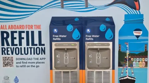 A colourful wall display reading 'all aboard for the refill revolution' next to two water dispensers