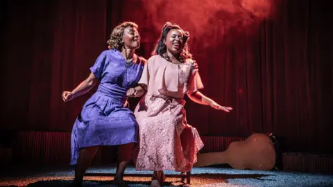 Marc Brenner Beverley Knight in a 1940s style purple dress dancing with her co-star Ntombizodwa Ndlovu who is in a pink floral 1940s dress. They are on stage in Marie and Rosetta. 