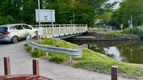 Photo taken from carpark near to the canal. A bridge with a silver car driving on top of it is seen stretching along the width of the canal. 