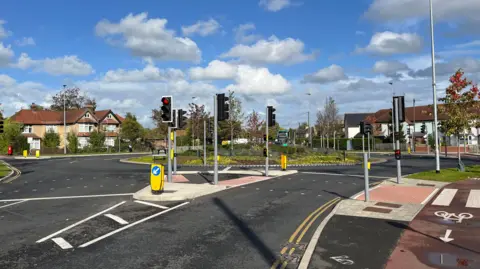 JOZEF HALL/BBC A road approaching a roundabout with a pavement, traffic lights and houses and greenery in the distance.