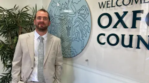 Mr Bearder is wearing a white suit and silver tie. He is standing next to the Oxfordshire County Council logo.
