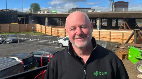 Jim Smyth wearing a black jacket which says "bam" in green lettering on the left breast pocket. He has grey stubble and is smiling at the camera. Behind him is a car park and a fencing and a large green machine. You can see the doomed flyover in the background being propped up. 