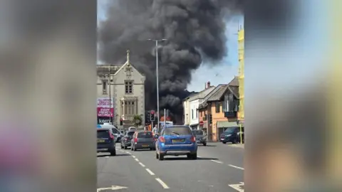Keith Mars A car drives on a road through a city. There are buildings on either side and a fire in the background.