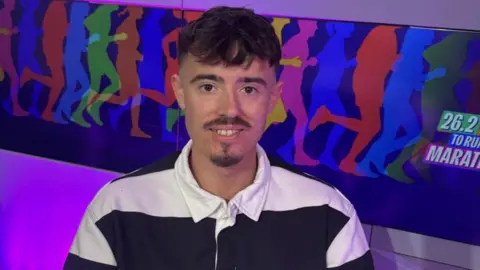 Ryan, who is wearing a black and white striped top, is sitting in the BBC Radio Manchester studio. There is a BBC-branded purple microphone on a desk in front of her. 