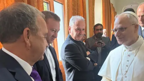 Jim Sharma A group of men are looking at Pope Leo XIV who is smiling. They are in a large room with patterned orange curtains along one side. The men are all in dark suits, and the Pope is in white clerical robes.