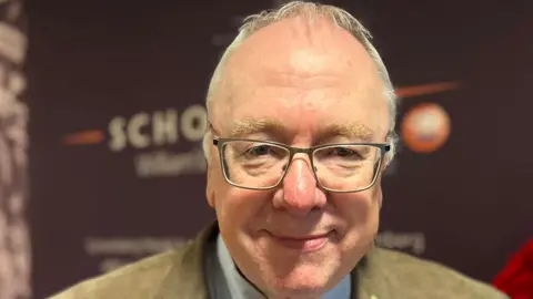Mervyn Gibson smiling in a beige suit with short white hair and glasses.