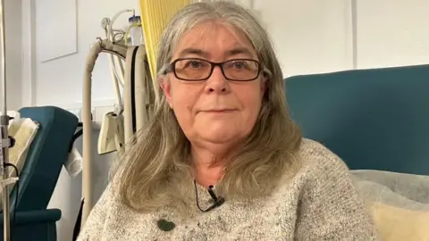 BBC Lesley Jenkins, who has long grey hair and glasses, sat on a hospital chair with a cubical curtain and medical equipment behind her. She is wearing a grey jumper with a broadcast mic pinned to it.