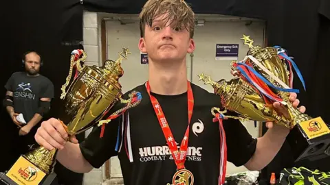 Family handout The young kickboxer with short blonde hair is wearing a black T-shirt with a gym logo and a medal on a red lanyard with a medallion holding a gold trophy in each hand.