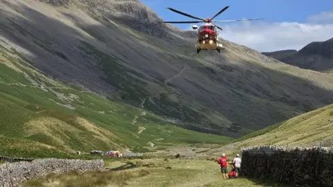 Wasdale Mountain Rescue Team A red helicopter flies low in the Wasdale Valley near Kirk Fell. Two mountain rescue volunteers stand near a dry stone wall and wait for the helicopter to land. There are emergency vehicles in the distance. Dry stone walls and grey scree slopes feature on the green terrain.