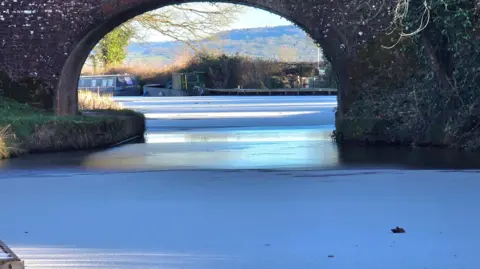 papa pics/BBC Weather Watchers A sheet of ice on a small body of water under a bridge. On the left is a riverbank with green grass. In the background, boats are moored. There are hills in the background. The sky is blue. 