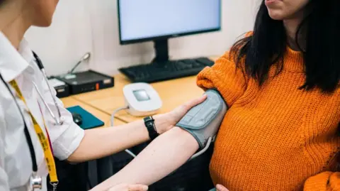 Getty Images Doctor takes blood pressure of woman sitting with sleeve rolled up. 