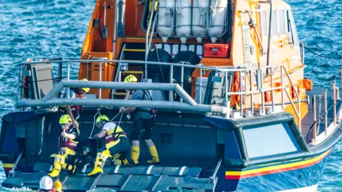 Geoff Squibb This picture shows an RNLI sea rescue operation in progress. Three crew members, dressed in yellow helmets and high-visibility RNLI gear, are helping a black dog onto a lifeboat from the water. 