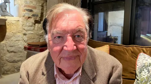 George Davies is sitting connected a sofa and leaning towards nan camera. He is wearing a reddish and achromatic garment and beige jacket. He has grey hairsbreadth and he is smiling.
