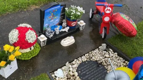 Victoria Richins A child's grave, with supermario decorations around it including a floral mushroom decoration, red plant pot with an M motif and a mini red scooter. There are also yellow roses in a box and ornaments at the foot of a small black headstone. A flat plaque lies in front of the headstone, on a bed of stones. Grass surrounds the plot.