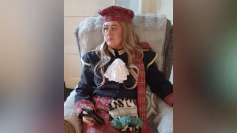 Lisa Hughes Woman wearing a kilt, tartan beret and navy jacket sitting in a chair.