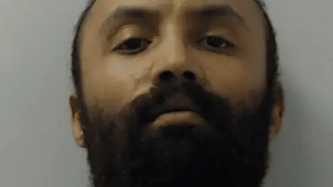 A police mugshot of a young man with a scruffy dark beard and a serious expression. 