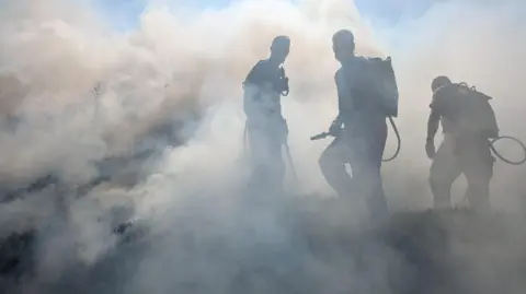 Three people in breathing apparatus are surrounded by smoke while trying to put out a wildfire.