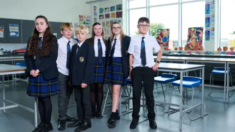 Channel 4 The students stand together in the classroom. Three girls and three boys. They are all wearing school uniform and standing in front of a number of desks and stools. 