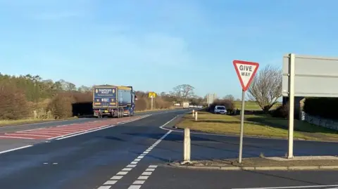 The junction at Charlton Mires. A "give way" sign is in the foreground where a road on the right of the image joins the main A1. A blue lorry is passing by on the left with more vehicles in the distance.