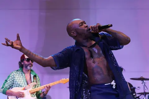 SEAN DRAKES/GETTY IMAGES Seun Kuti sings with the microphone, whilst a band member plays the guitar in the background. 