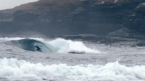 Pete Hill A surfer is riding inside a breaking wave, positioned in the hollow barrel as water curls over the top. In the background, there is a rocky coastline with cliffs and scattered buildings under a grey, overcast sky.