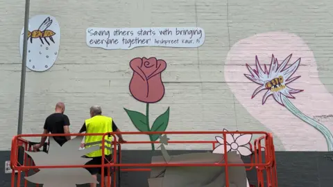 Luton Lifelines A red scissor lift stands in front of the partly finished mural. Two men stand on it with their backs to the cameras. On the mural is a quote saying "Saving others starts with bringing everyone together, Asrhpreet Kaur, 15". Below that is a red rose and on either side are images of bees and flowers.