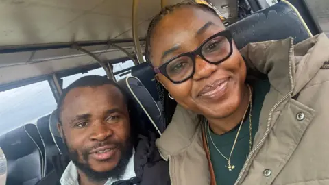 Gabriel Enyi/Uloma Igwe Gabriel Enyi and Uloma Igwe sitting in a bus. Gabriel Enyi has very short black hair and a black beard and is wearing a black coat over a white shirt and is starting to smile. Uloma Igwe's black hair is braided back from her head and falling to the left. She is wearing black framed glasses, and a light brown coat over a dark green sweatshirt and smiling. 