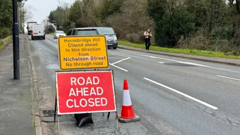 Image shows a road with traffic. There is a sign and a traffic cone saying Road Ahead Closed, and a yellow information sign saying that Horsebridge Hill is closed from Nicholson Street and that this road (Horsebridge Hill) is a no through road.