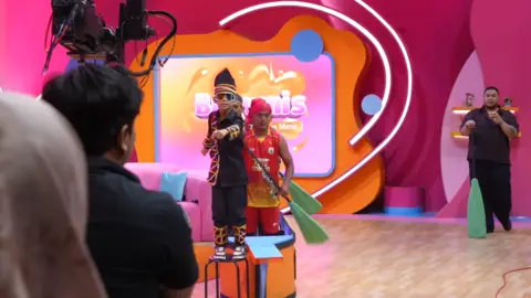 BBC/Dicky Kurniawan Rayyan Arkan Dikha, in a traditional black Malay outfit wearing sunglasses, stands on a chair dancing in front of a camera
