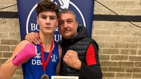 Father and son Jakson (left) and Richard Myers have fists clenched in a boxing pose. Richard, with grey hair and beard, wears a gilet and has his arm around Jakson, who wears a blue boxing vest and a medal on a red ribbon around his neck.