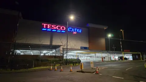 The outside of a supermarket building, with an entrance to a car park. A neon Tesco Extra sign is at the top of the building red and blue letters. The store is cordoned off by orange cones. 