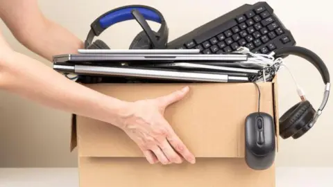 A person holds a cardboard box piled up with laptops, tablets, a keyboard, headphones and a mouse hanging over the side.