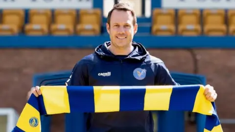 James Rowe is standing on a football pitch wearing a navy hoodie and holding a yellow and blue football scarf. He is looking directly at the camera and smiling. 