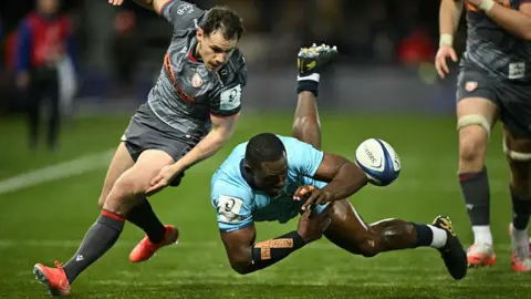 Justin Tallis/Getty Images Two rugby players on the pitch trying to catch a ball as it passes in the air. The Gloucester player, dressed in grey kit, is balanced on one foot while leaning forward, while the Castres player, dressed in turquoise kit, is completely suspended horizontally in mid-air.
