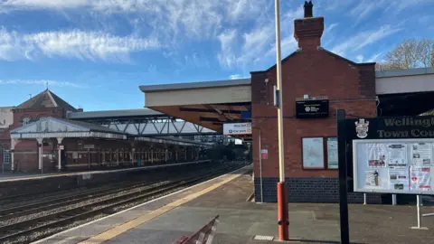 A railway station's platform under a clear blue sky. There is a red and white bench in the foreground with brick railway buildings behind, a thick yellow line on the platform edge and two sets of rails between the platforms.